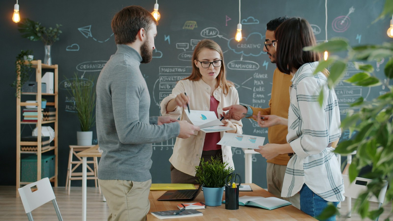 Four coworkers stand around a desk reviewing papers in front of a chalkboard