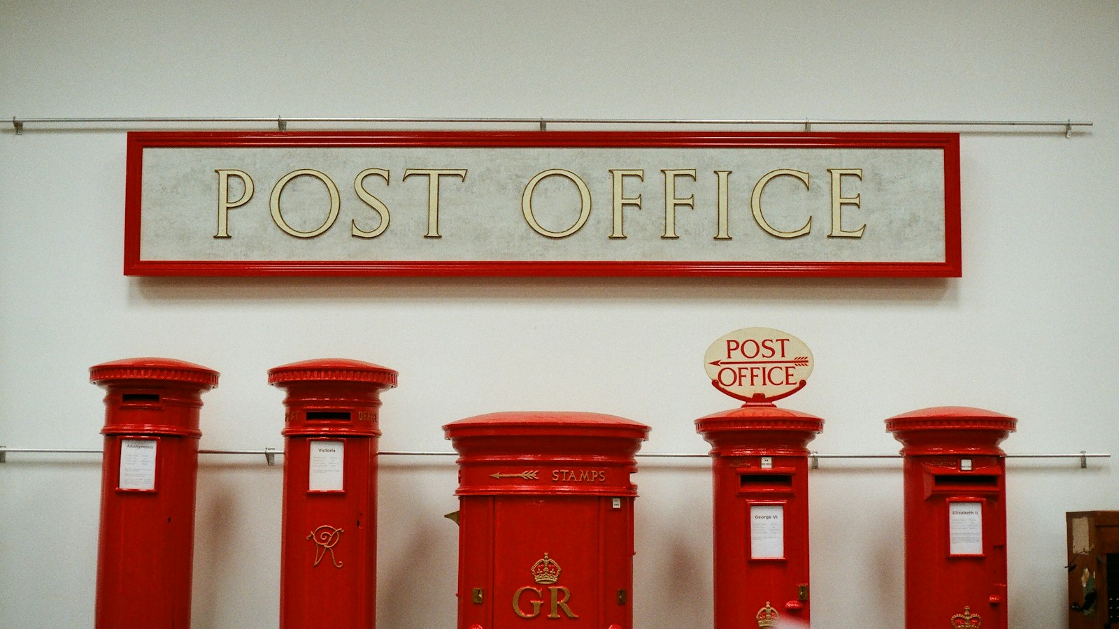Row of red post boxes beneath a post office sign on a white wall