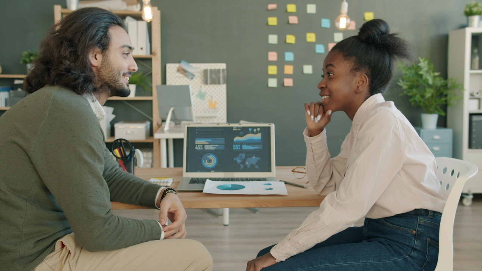 Two colleagues talking at a shared desk with a laptop in a bright office