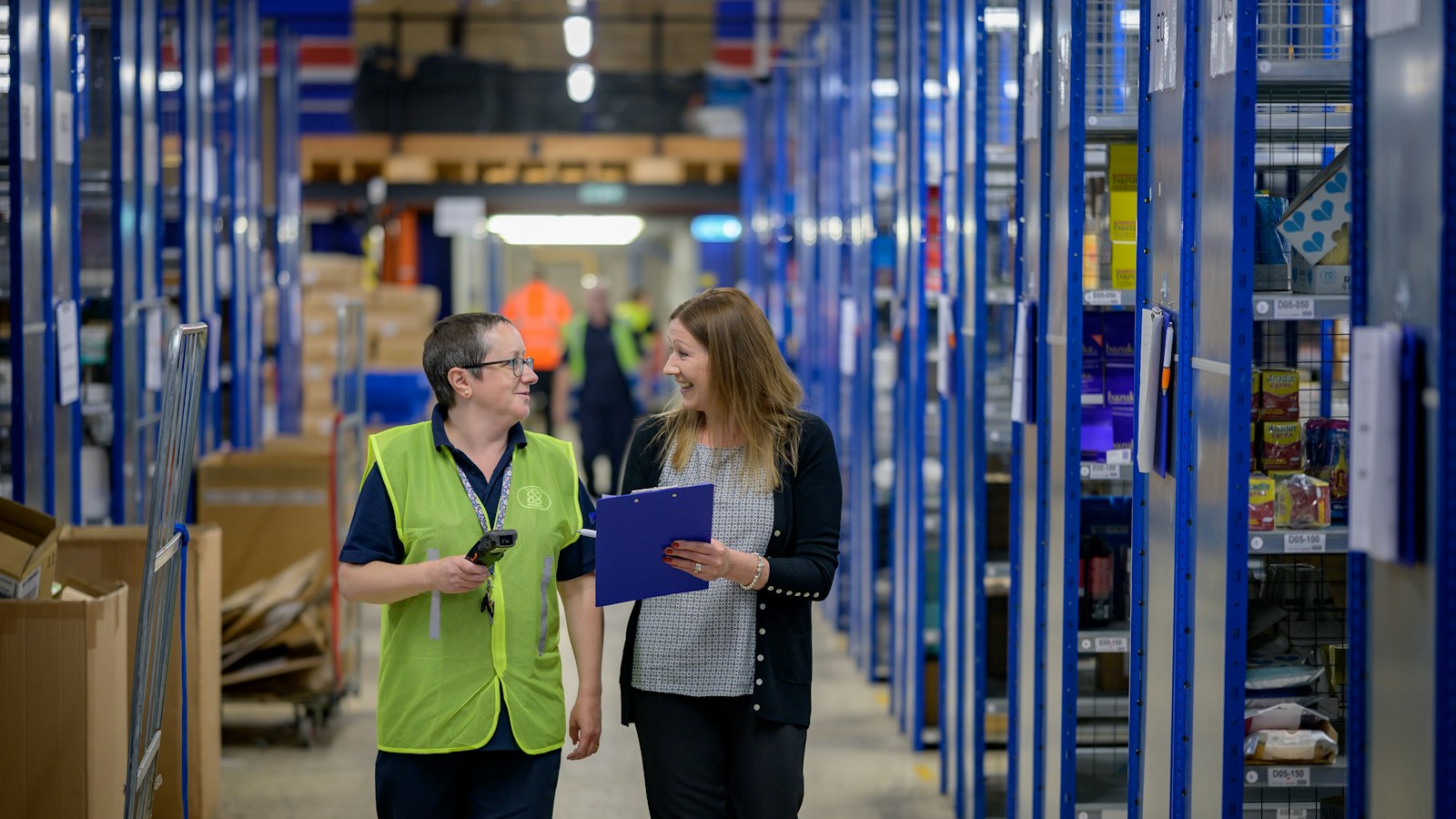 Two warehouse staff walk an aisle between blue shelving while one holds a clipboard