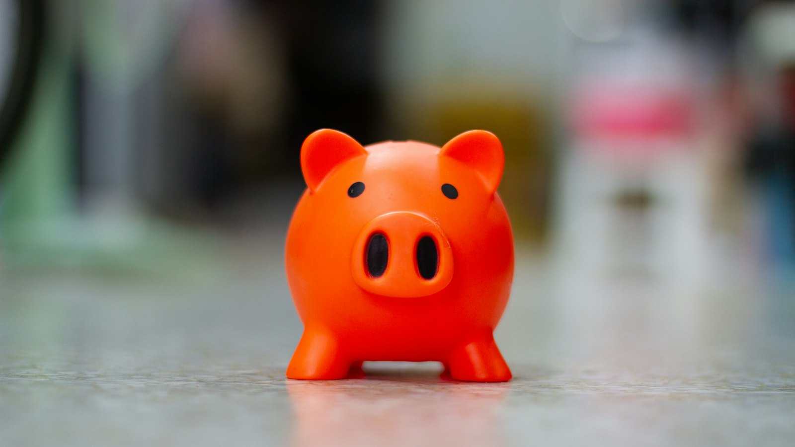 A small orange piggy bank sits on a pale floor with a blurred background