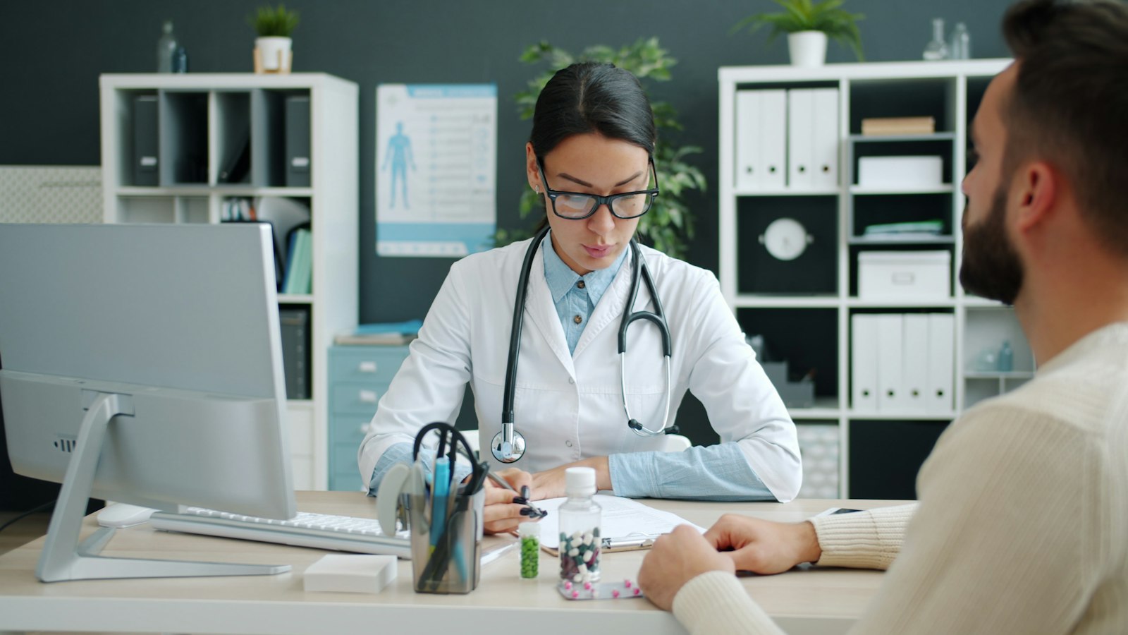 Doctor in a white coat writes notes while talking with a patient at her desk