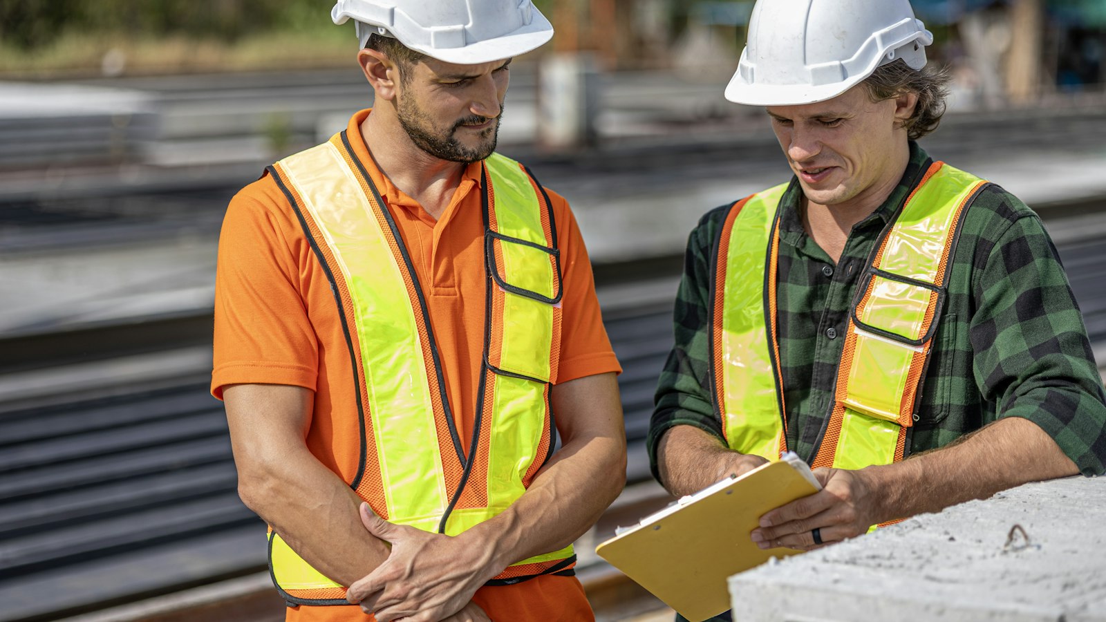 Two workers in hard hats and safety vests look at a clipboard outdoors