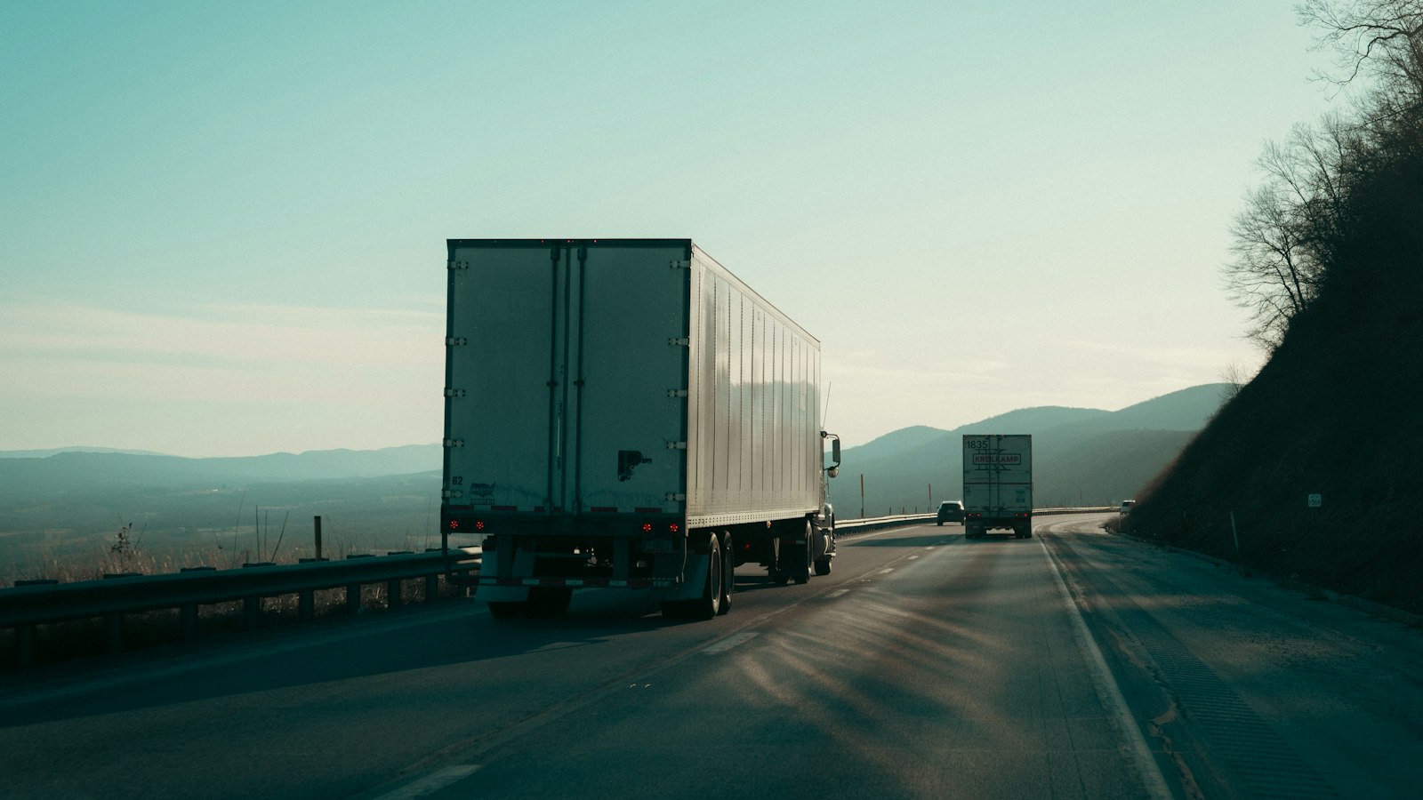 Two semi trucks driving on a highway toward distant hills at dusk