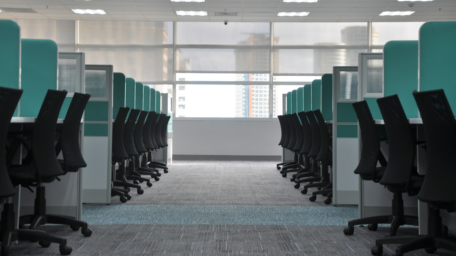 Rows of empty black office chairs at cubicles along a long open office aisle
