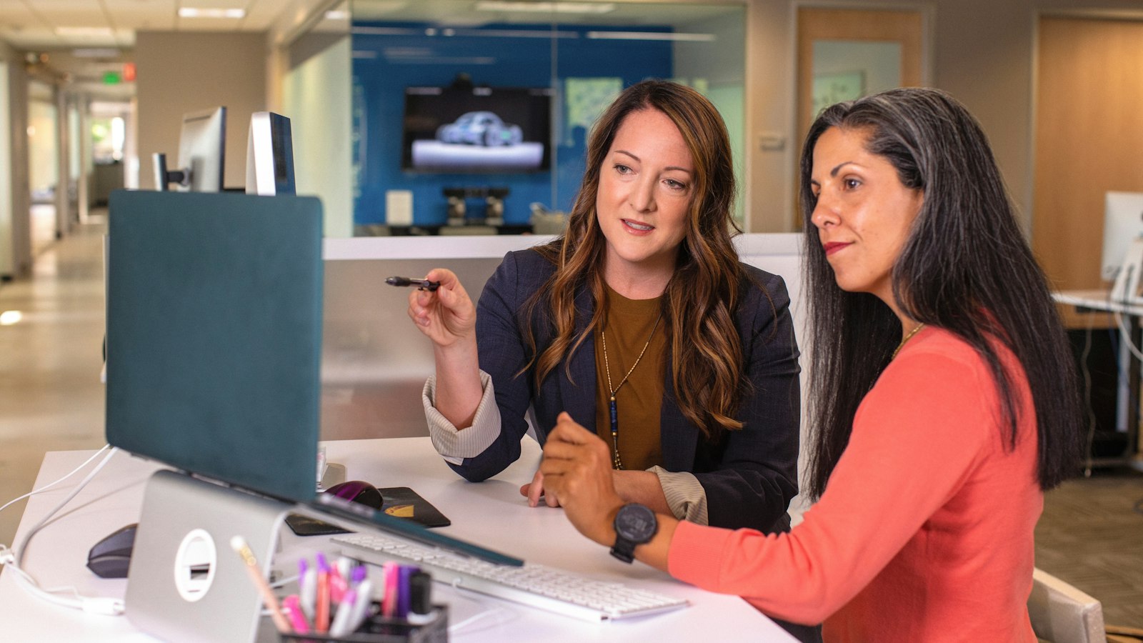 Two women sit at a desk in an office and look at a computer screen together
