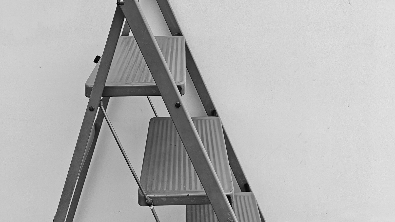 A black and white photo of a metal step ladder leaning against a plain wall