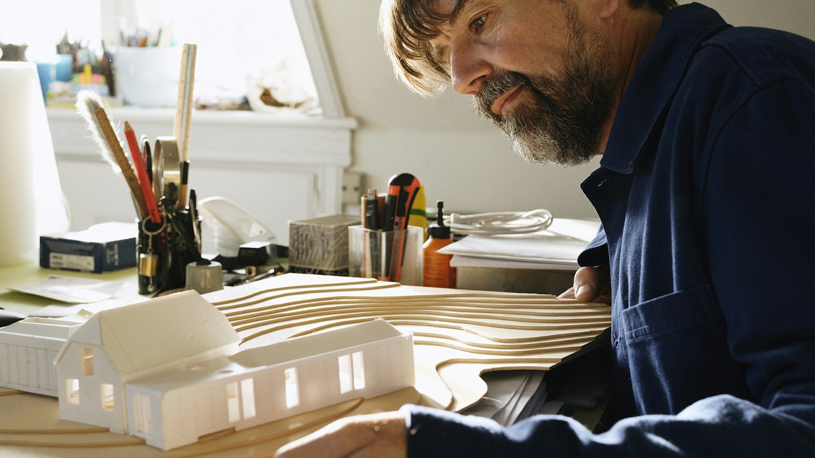 A bearded man leans over a small white architectural house model on a desk
