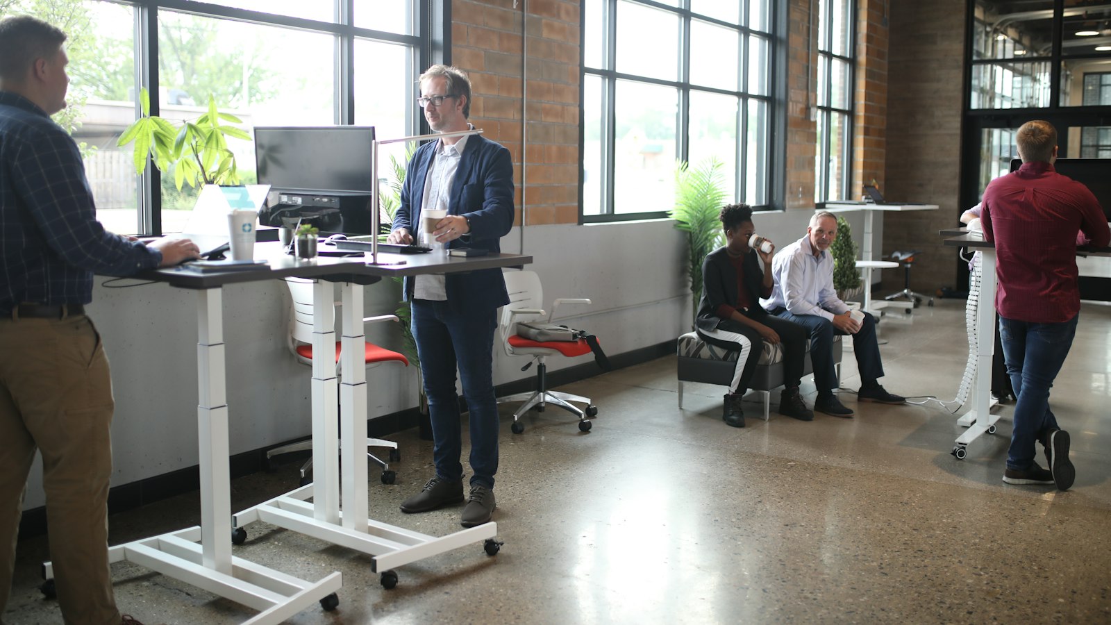 Colleagues work at standing desks and talk in a bright open office with brick walls