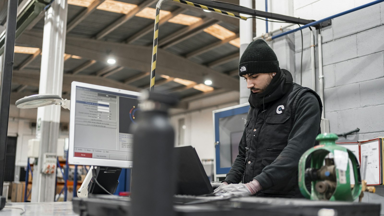 A man in a black beanie works at a computer in an industrial workshop