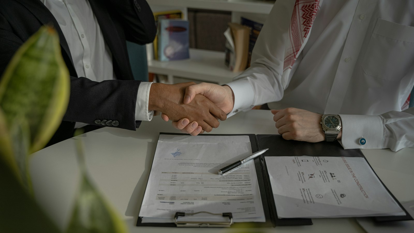 Two people in business clothes shake hands over a folder of documents and a pen