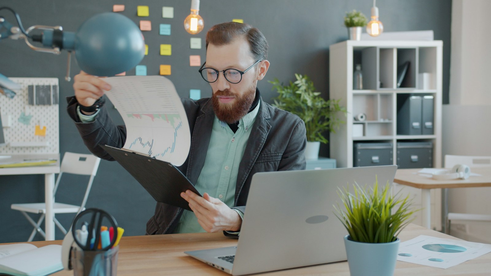 A bearded man in glasses holds up a printed chart beside his laptop