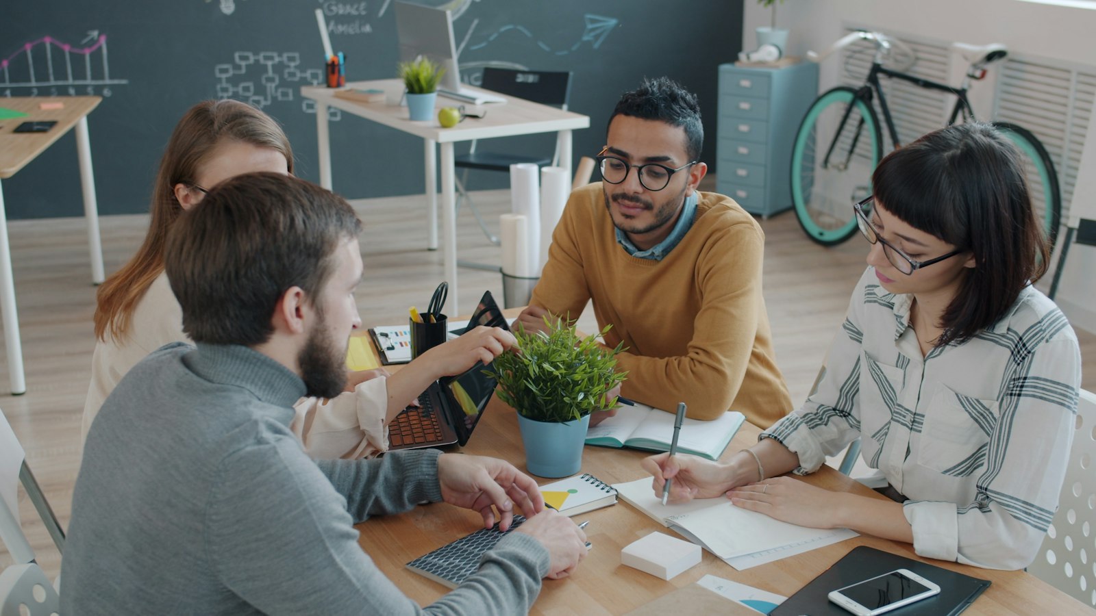 Four colleagues talk around a wooden table with laptops and notebooks in a bright office